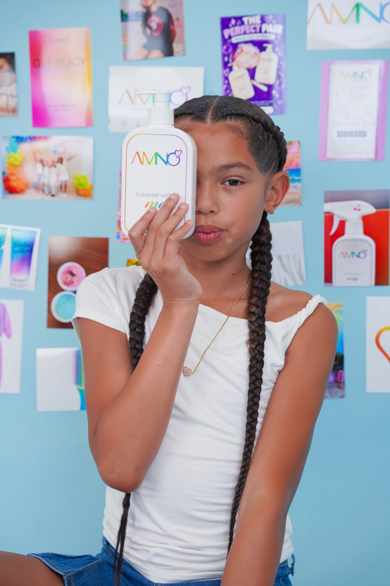 A girl with long braids holding the Cleanse With Me bottle over one eye in front of a wall of AMNO photos