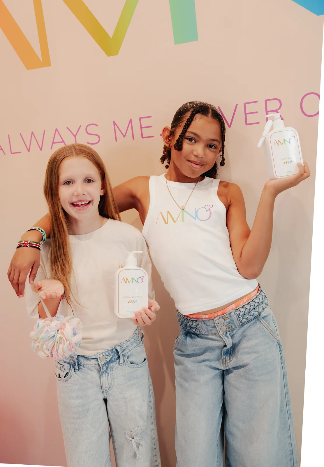 Two girls in AMNO tees holding Cleanse and Care bottles in front of the Always Me, Never Others wall