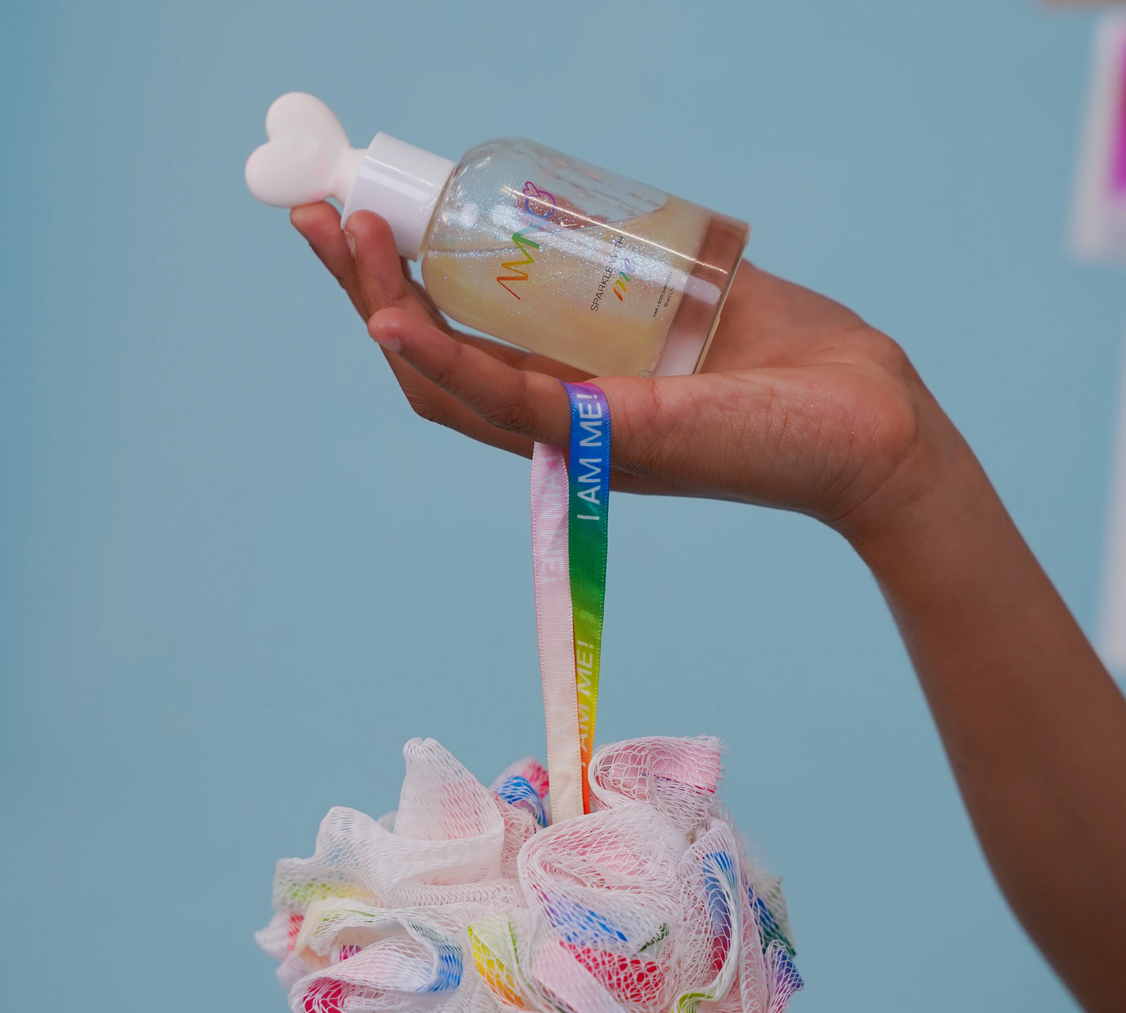 A hand holding the Sparkle With Me bottle with a rainbow loofah hanging from an “I AM ME!” ribbon