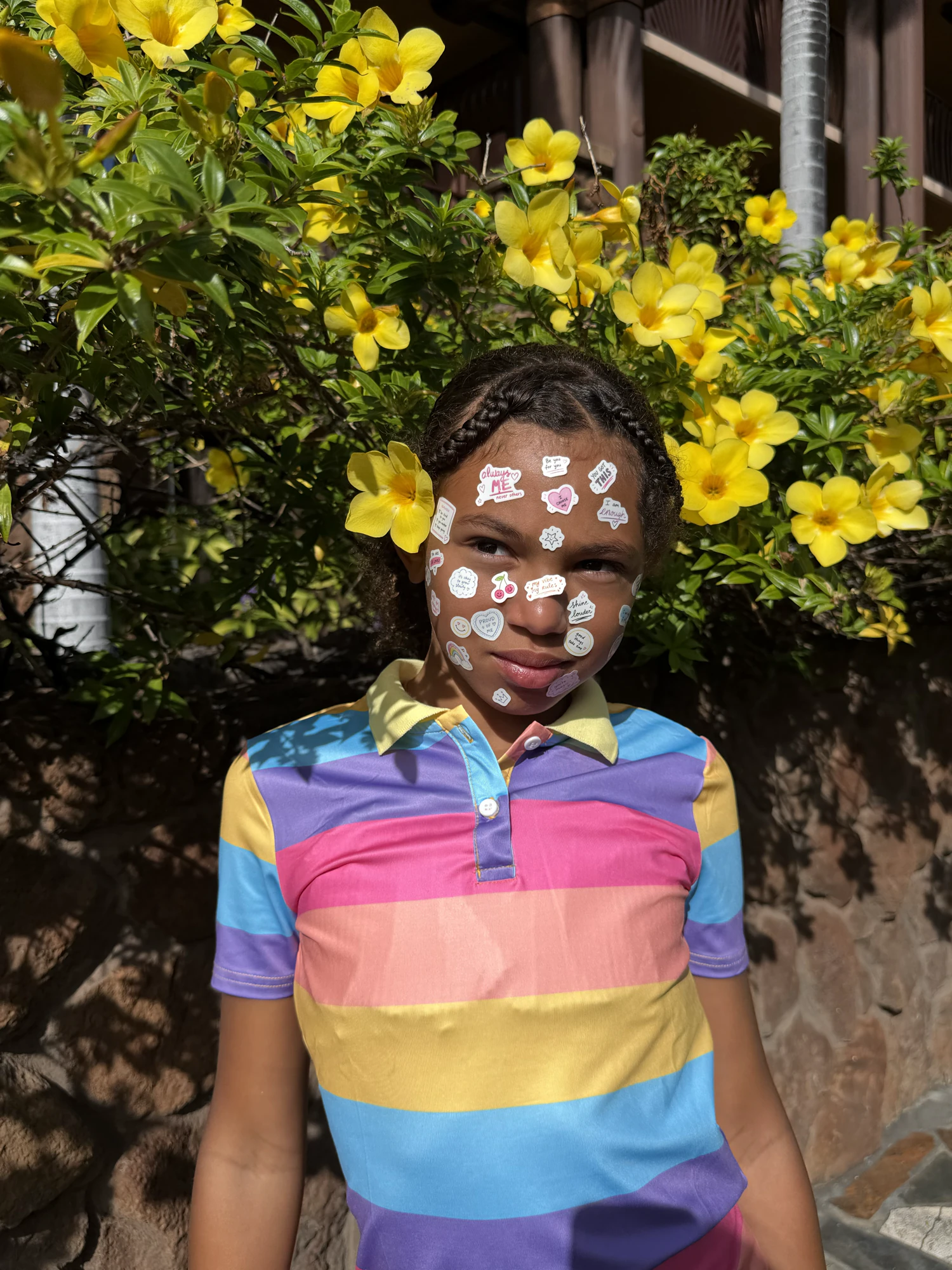 A girl in a rainbow striped polo with affirmation stickers on her face, standing in front of yellow flowers