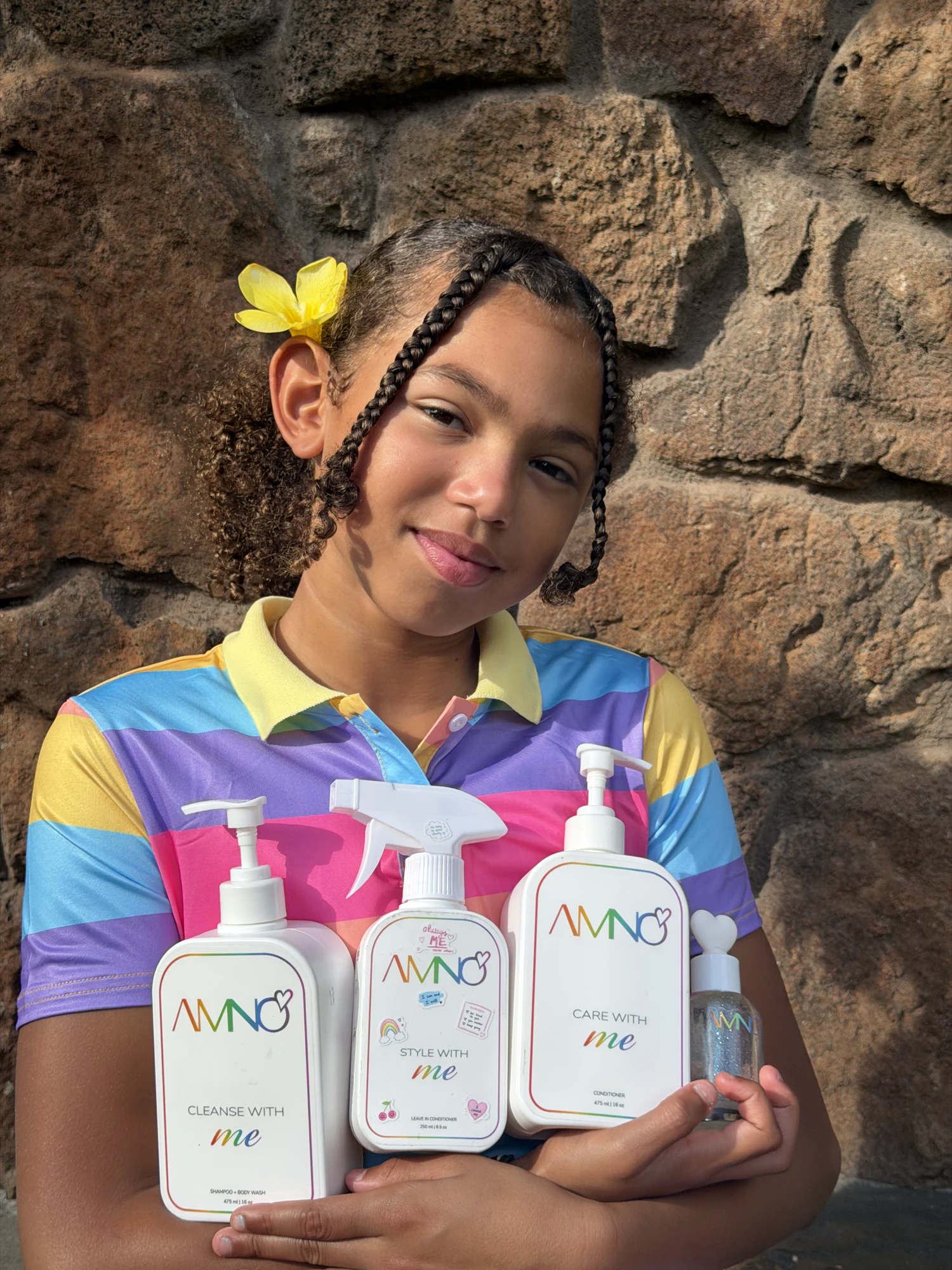 A girl in a rainbow polo holding all four AMNO products in front of a stone wall